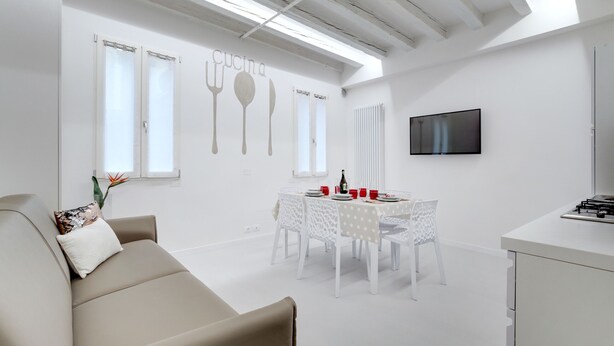 A modern, minimalist living room with a sofa, a dining table set for four, and a kitchen area with a TV. - Bellini Design by Wonderful Italy (Venecia)