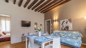 Un comedor con una mesa dispuesta para cuatro, sillas blancas, un sofá, un televisor de pantalla plana montado en la pared y una puerta que da a otra habitación.