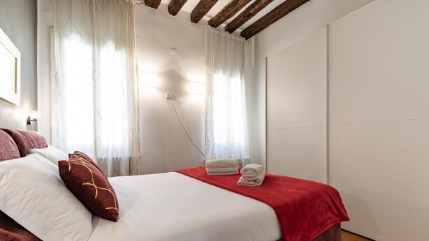 Una cama bien tendida con una colcha roja, un cabecero de madera y una lámpara fijada a la pared.