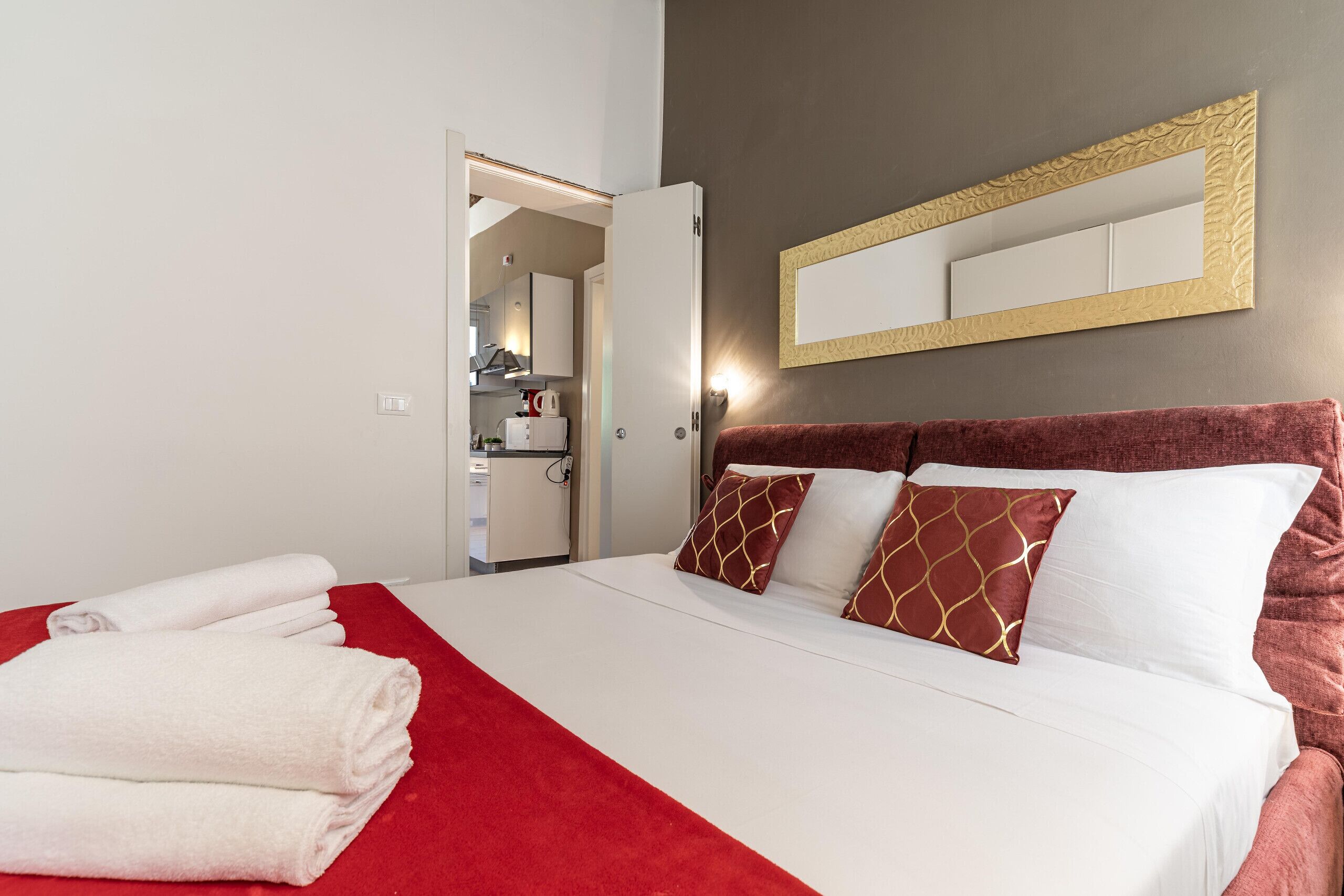 Une chambre d’hôtel avec un lit, un plaid rouge, des serviettes blanches, une tête de lit rouge et un miroir avec un cadre doré.