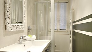 A modern bathroom with a white vanity, a large mirror with ornate detailing, a shower with a glass door, and a window providing natural light. - Ca' Forner by Wonderful Italy (Venecia)