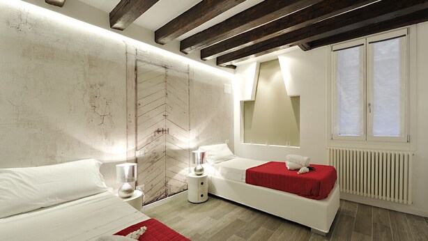 A modern bedroom with a bed, a sofa, a red accent wall, a large mirror, and a window with blinds. - Ca' Forner by Wonderful Italy (Venecia)