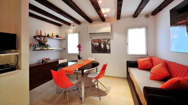 A dining area with a round table, red chairs, and a sofa with orange cushions. - Creme Caramel Room by Wonderful Italy (Venecia)