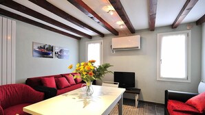 Ein Zimmer mit einem roten Sofa, einem Tisch mit Blumenvase, einem Fernseher und einem Fenster mit Klimaanlage.