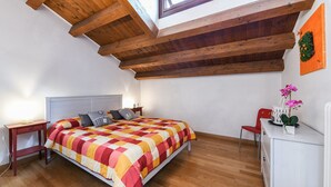 Een slaapkamer met een houten plafond, een bed met een kleurrijke sprei, een nachtkastje, een stoel en een klein tafeltje met een plant.