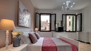 Ein Schlafzimmer mit Bett, Nachttischen, einer Lampe, einer Vase mit Blumen, einem Gemälde, einem Spiegel und Blick auf Gebäude durch ein offenes Fenster.