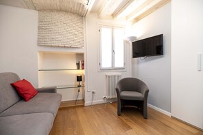 Ett modernt vardagsrum med en grå soffa, en röd kudde, en glashylla med böcker, en grå fåtölj, en platt-TV som är monterad på väggen och ett fönster med ett vitt element under.