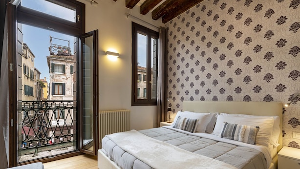 A bedroom with a bed, a nightstand, a window with a view of buildings, and a balcony with a railing. - Ca' Beccarie 2 by Wonderful Italy (Venecia)
