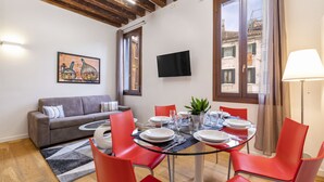 A dining area with a round glass table set for four, surrounded by red chairs, a grey sofa, and a flat-screen TV on the wall. - Ca' Beccarie 2 by Wonderful Italy (Venecia)