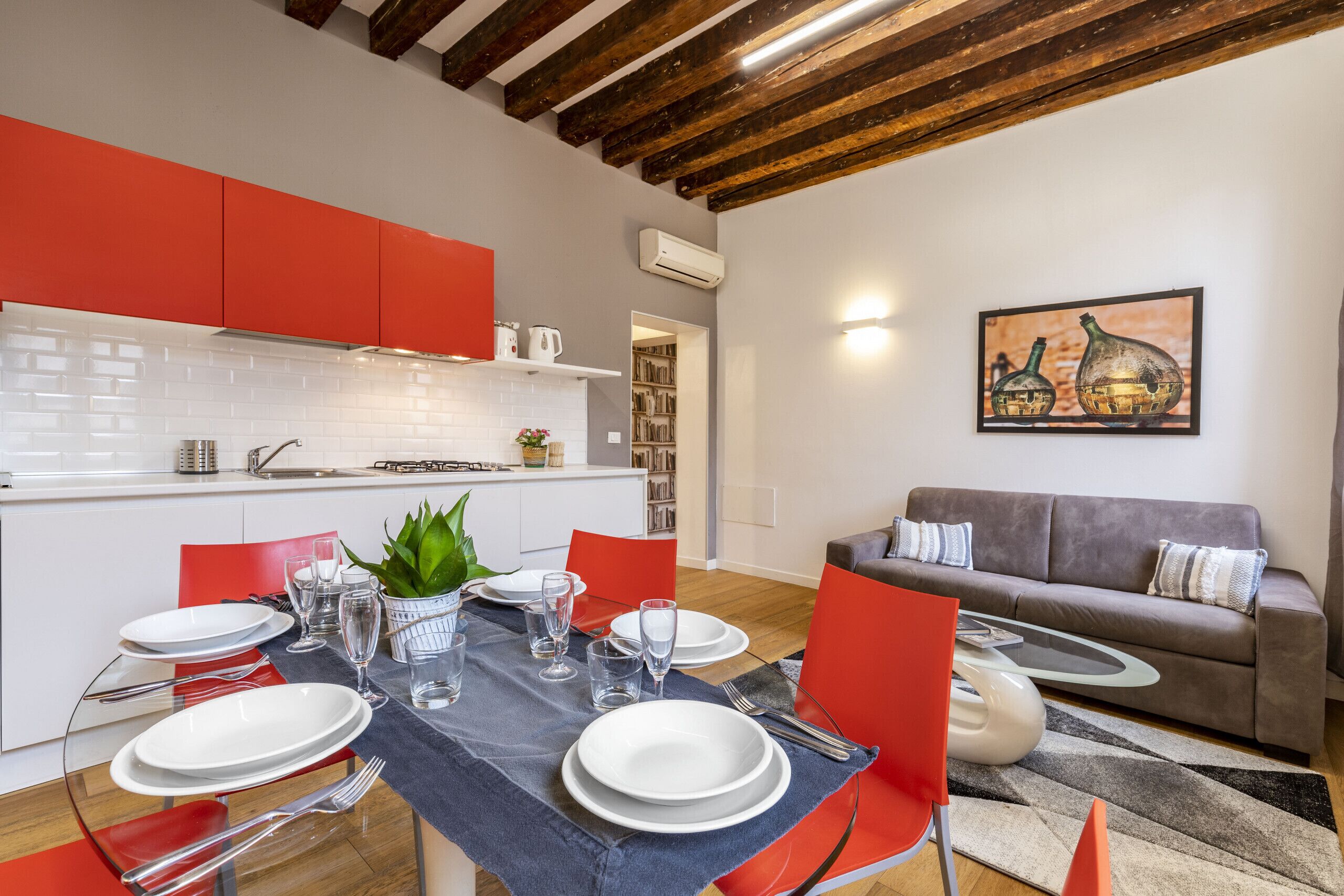 A modern kitchen with red cabinets, a dining area with a table set for four, and a living room with a sofa and a painting on the wall.