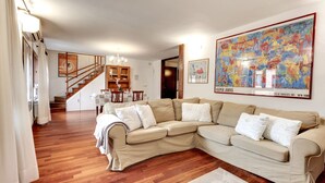 Ein geräumiges Wohnzimmer mit einem großen, beigefarbenen Ecksofa, Holzdielenboden, einer Treppe, die zu einer oberen Ebene führt, und einem farbenfrohen abstrakten Gemälde an der Wand.