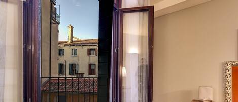 Una habitación con piso de madera, una mesita de noche de madera con una lámpara, una cama con colcha estampada y vista a un edificio con tejado de tejas a través de una puerta abierta.