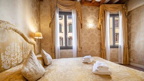 Habitación de hotel con una cama grande, dos ventanas con cortinas, una lámpara de pared y vista a un edificio a través de las ventanas.
