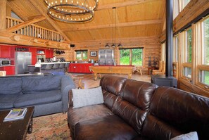 A log cabin interior with a living room, kitchen, and dining area, featuring wooden beams, large windows, and a fireplace.