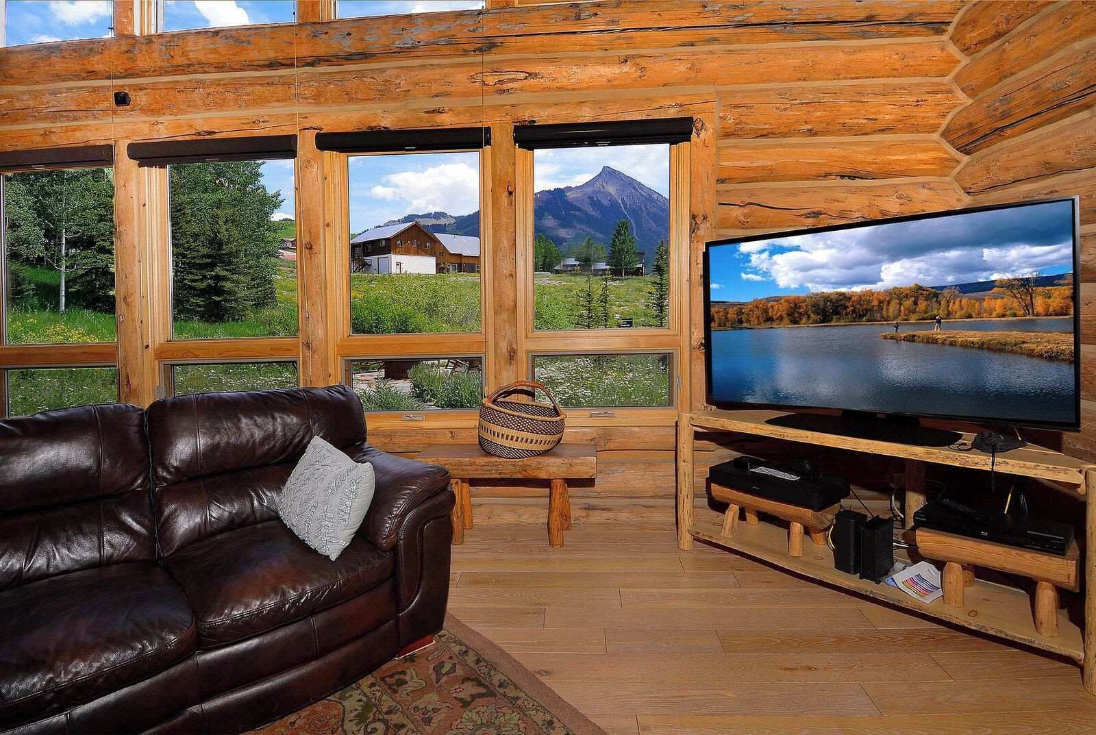 A wooden cabin interior with a leather couch, a television, and a large window offering a view of a mountain and a lake.