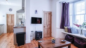 A compact living space with a wooden floor, a TV mounted on the wall, a window with purple curtains, and a small table with decorative items. - Sea Glass - 2 Bedroom Apartment - Manorbier (Tenby)