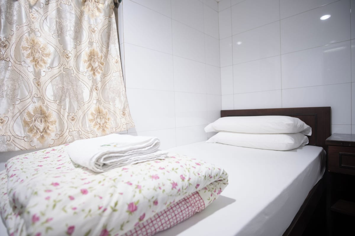 A neatly made bed with white linens and a floral-patterned blanket, a wooden headboard, and a white tiled wall.