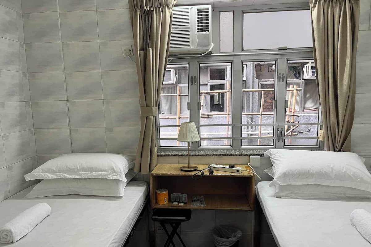 A small, clean room with two single beds, a desk with a lamp, and a window with curtains.