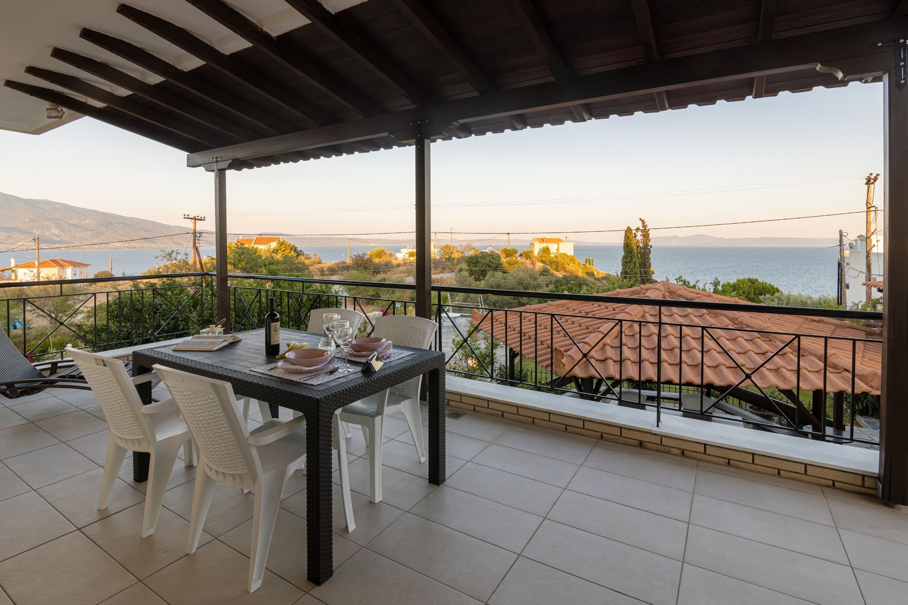 A covered outdoor dining area with a table set for two, overlooking a scenic view of the sea and hills.