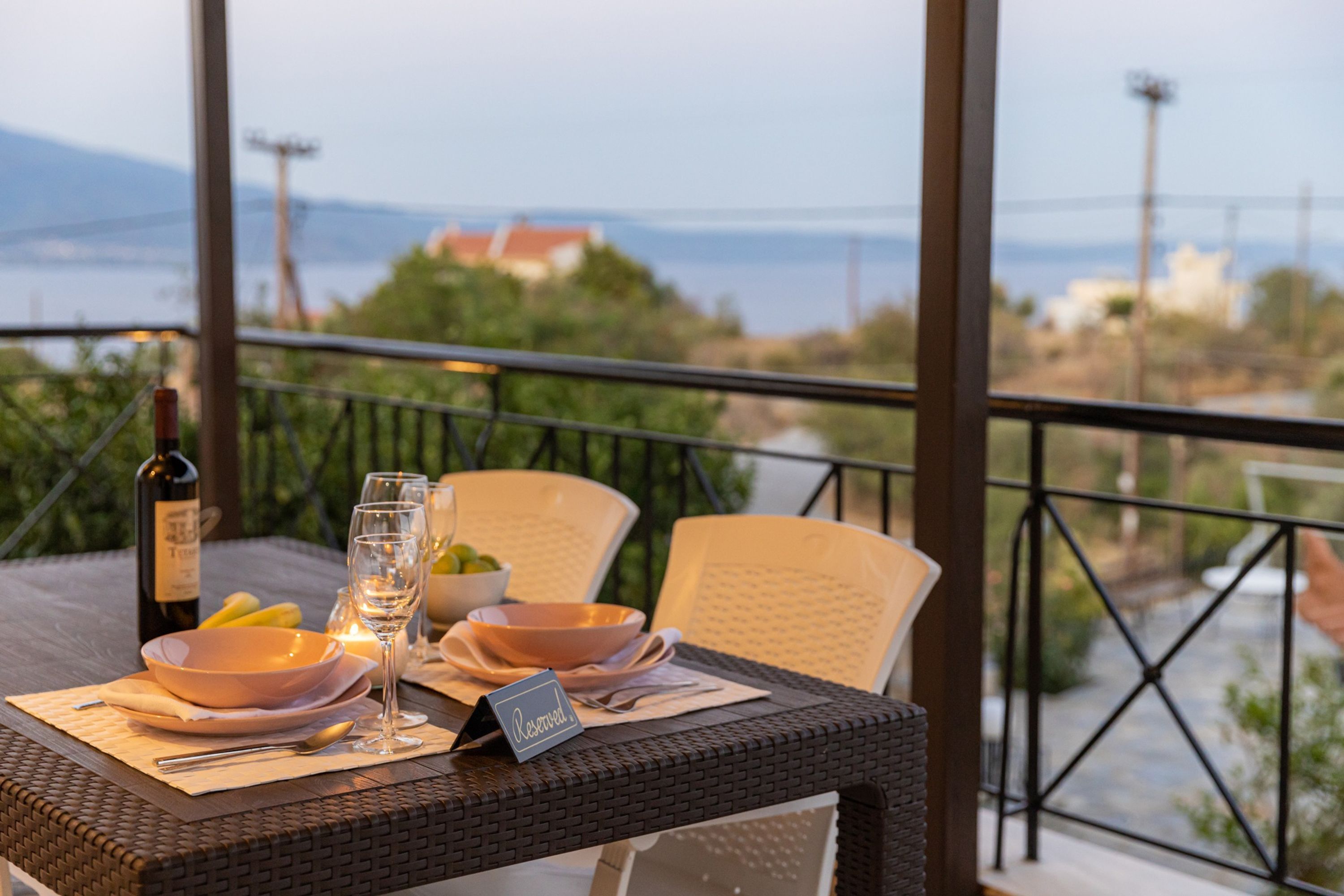 Une table à manger est dressée avec des assiettes, des verres et une bouteille, et offre une vue pittoresque sur un paysage verdoyant avec un bâtiment situé au loin.