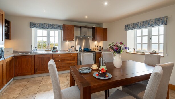 A modern kitchen with wooden cabinets, a dining table with chairs, and a view of the beach through large windows. - No. 9 The Dairy : Pet-Friendly 2‑Bed Barn Conversion Near Borthwen Beach | Managed by Anglesey Holid (Llanfaethlu)
