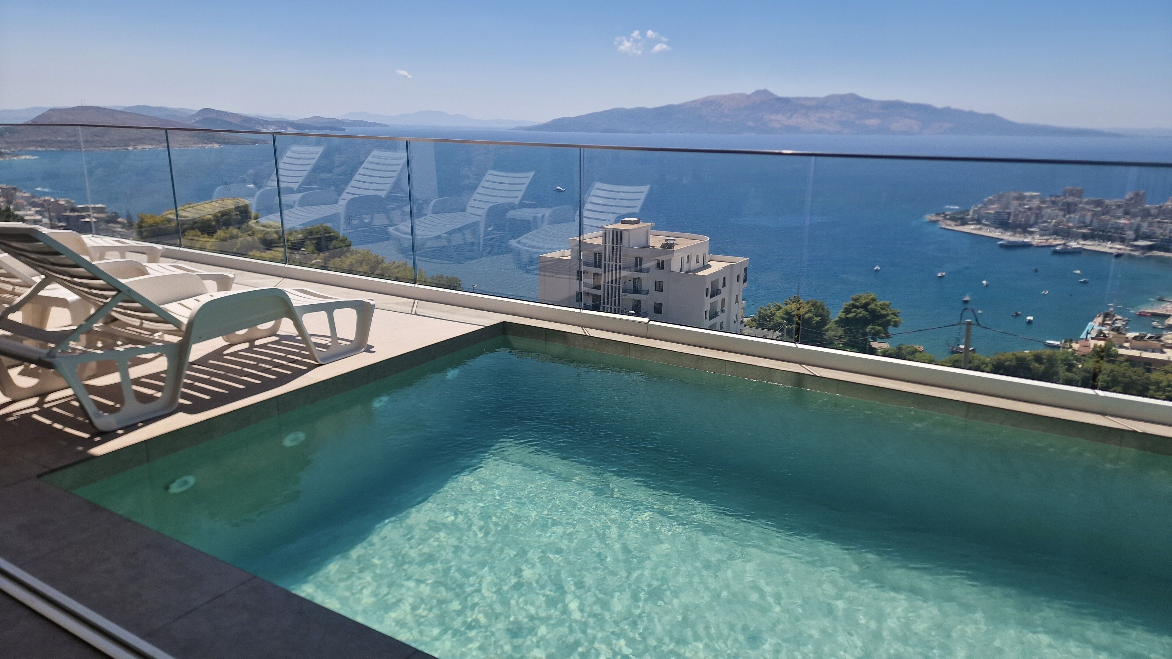 On peut admirer une piscine, des chaises longues, une balustrade en verre et une vue panoramique sur l’océan, avec des bateaux et une silhouette de ville qui se dessine à l’horizon.