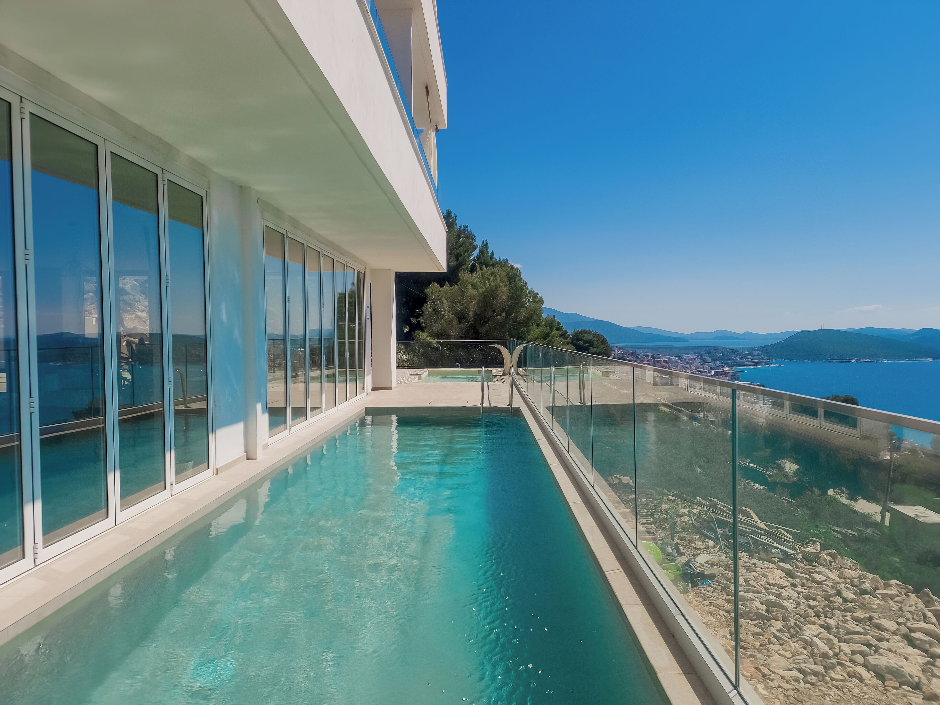 Un edificio moderno con balcón de vidrio que da a una piscina y una vista panorámica del mar y las montañas.
