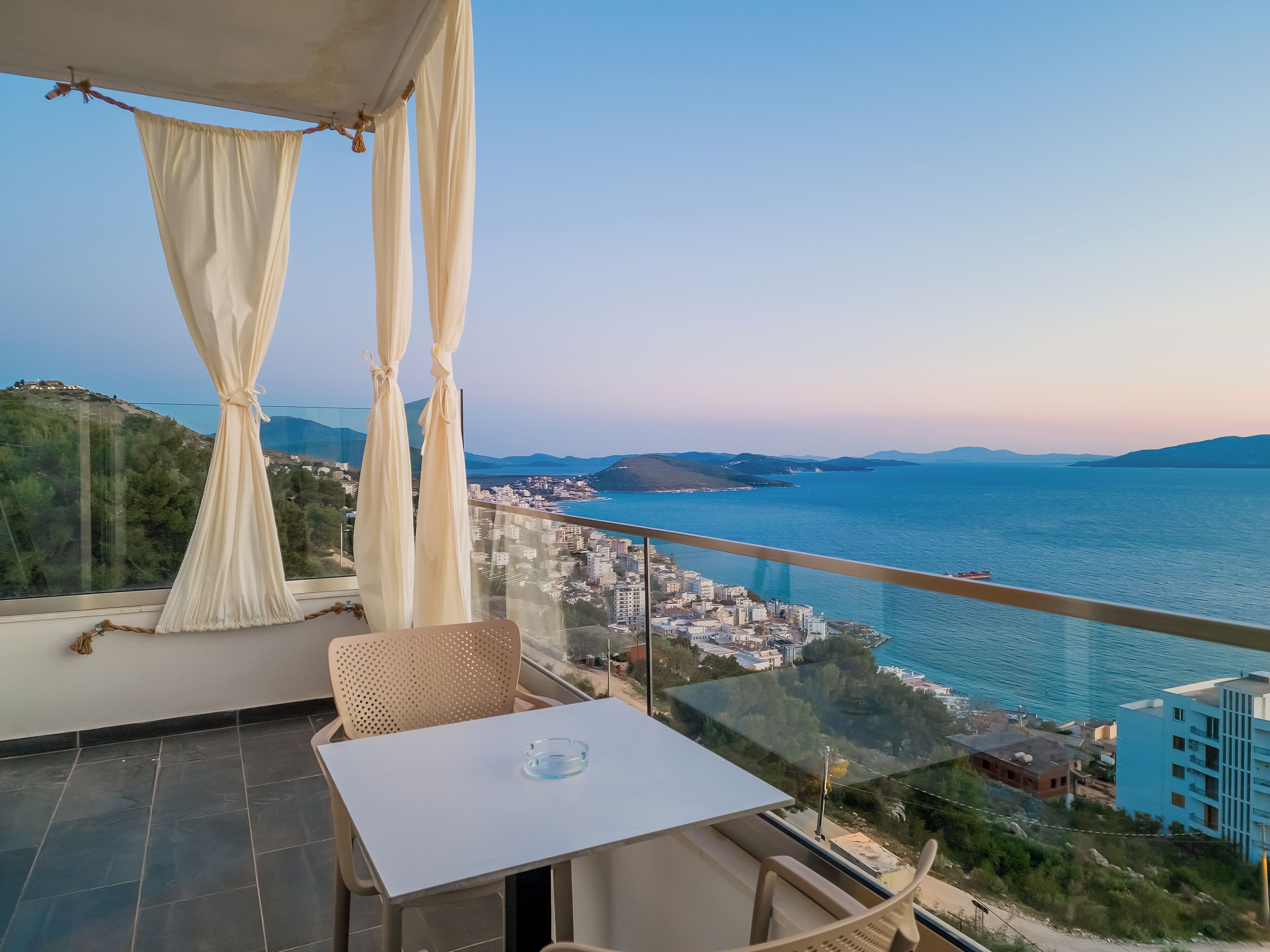 A balcony with a table and chairs, offering a view of a coastal city and the sea.