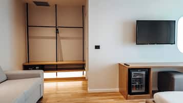 A modern hotel room with a wooden floor, a grey sofa, a bed, a TV mounted on the wall, and a built-in entertainment unit.
