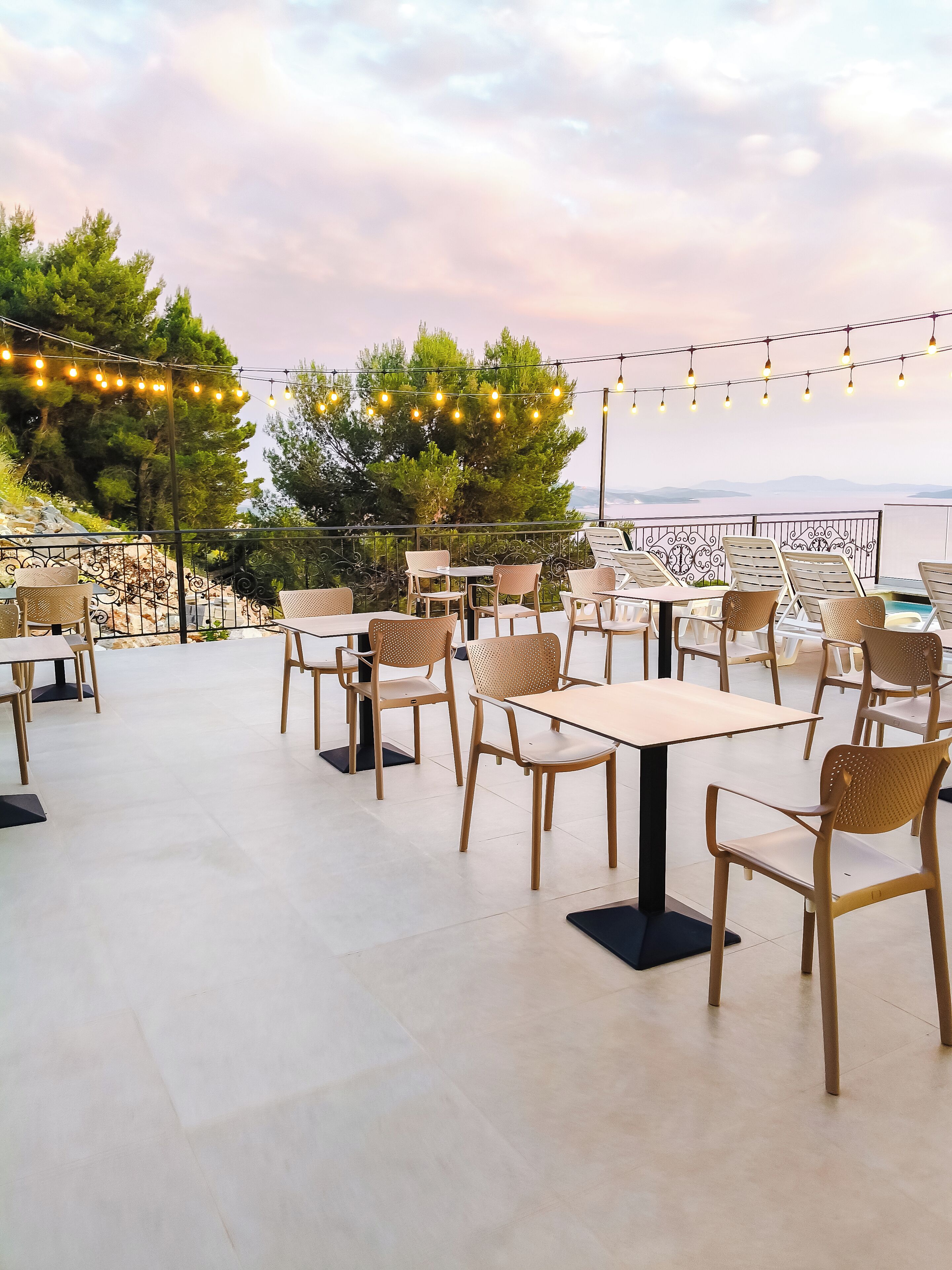An outdoor seating area with tables and chairs, string lights, and a view of the sea.