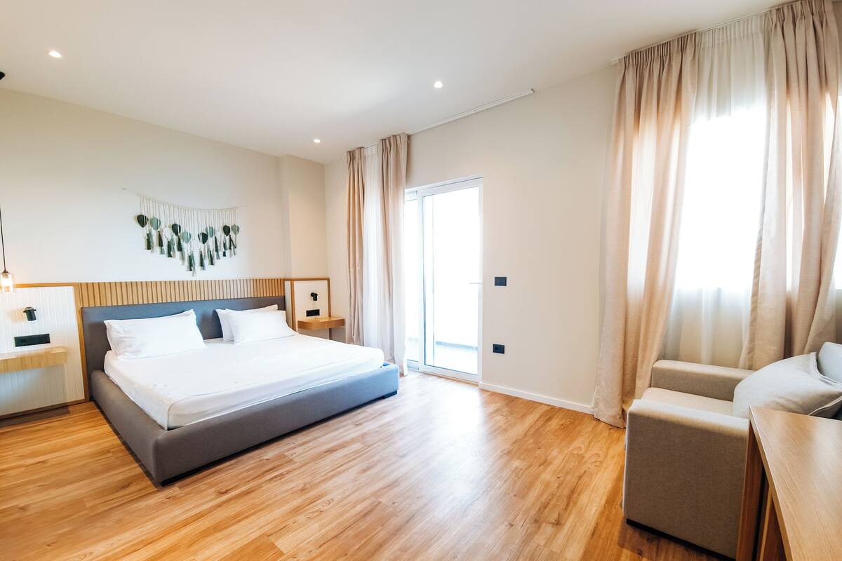 A modern hotel room with a large bed, wooden flooring, a seating area, and a view of the outdoors.