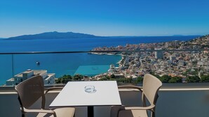 A table and chairs on a balcony overlooking a coastal city with a clear view of the sea and distant mountains. - Sunset Shoreline Saranda Hotel (Sarandë)