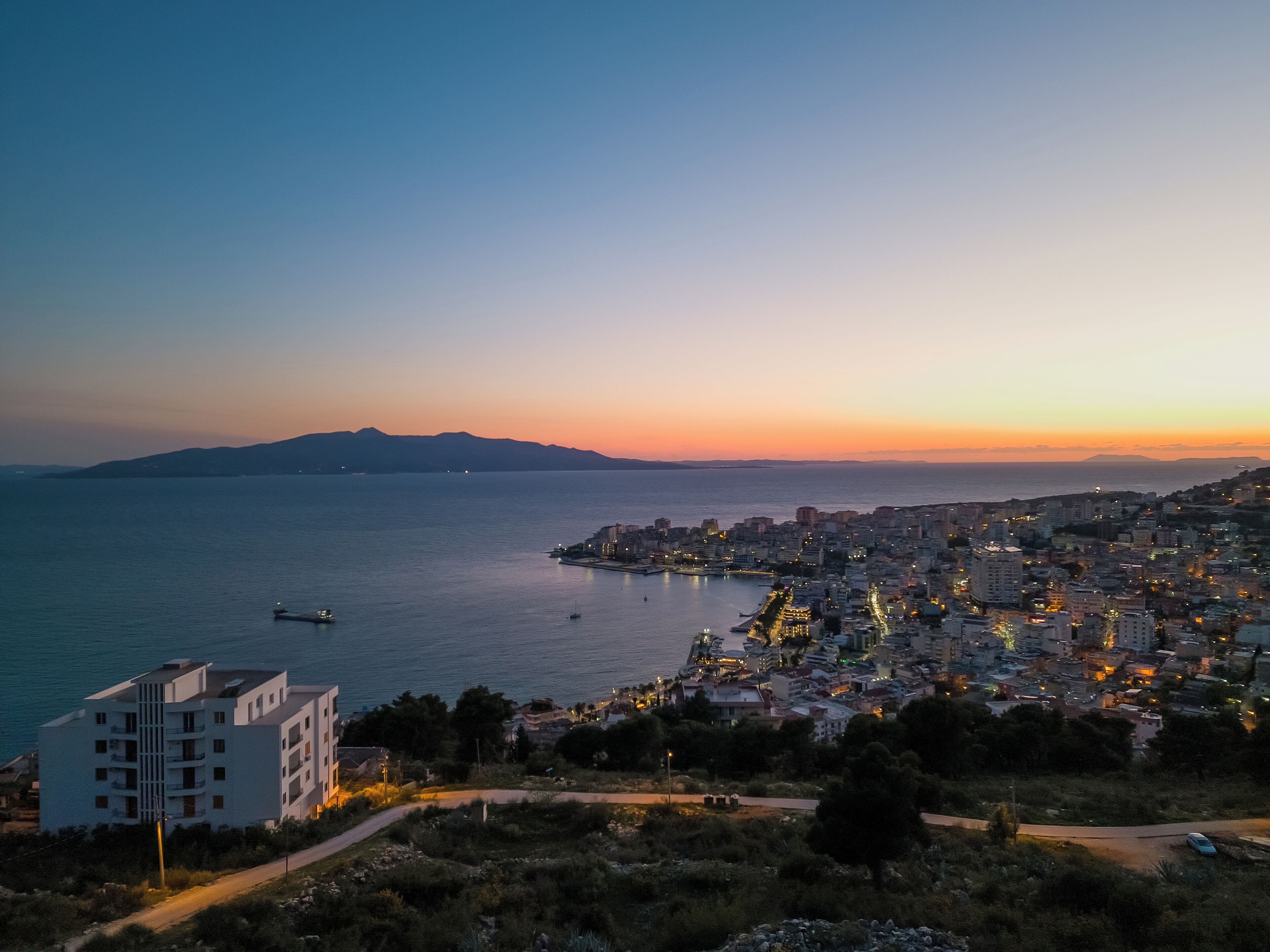 A coastal cityscape at dusk with buildings along the waterfront, a clear view of the sea, and a distant mountain range.