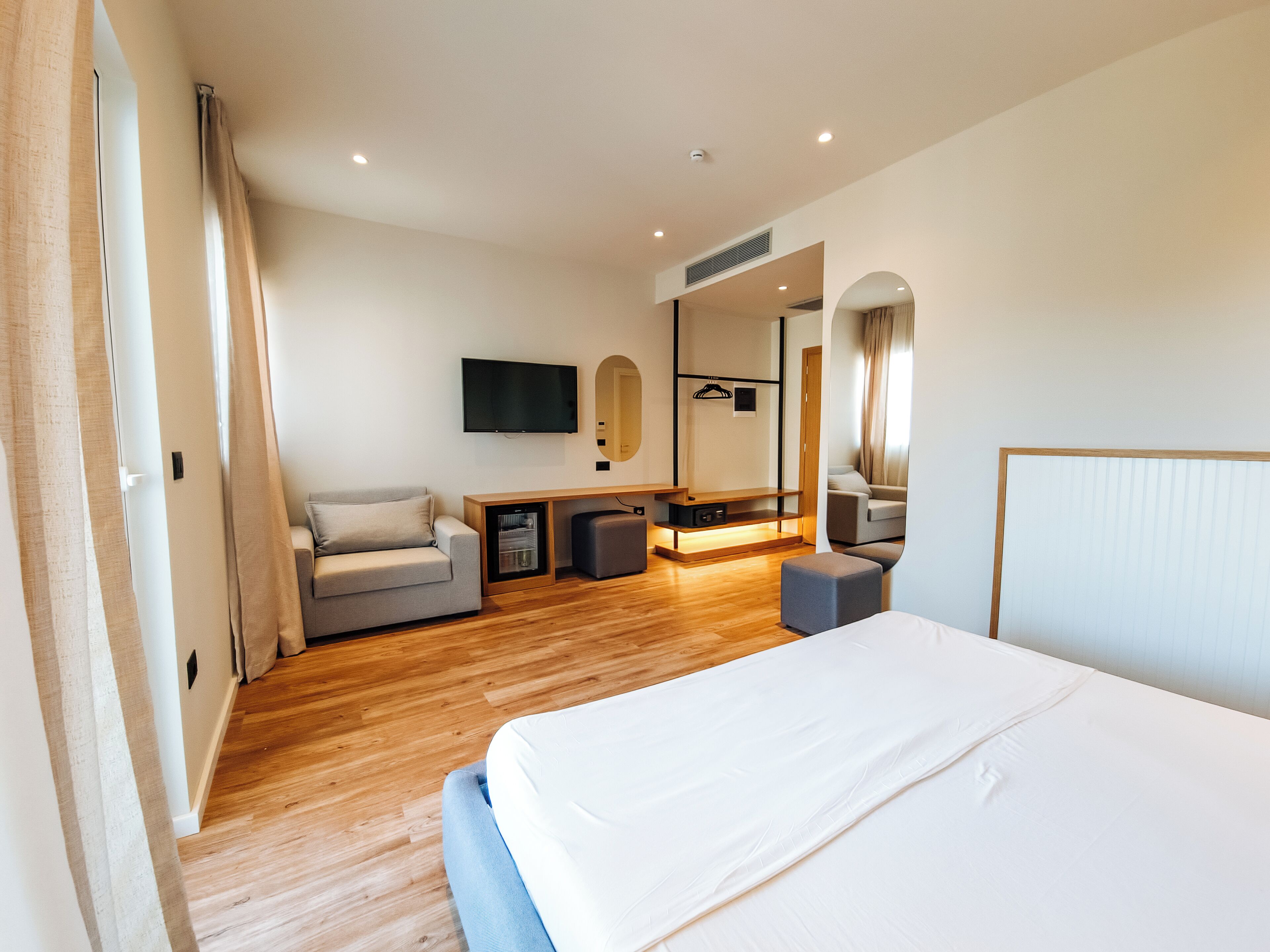 A modern hotel room with a large bed, a sofa, a TV, a mirror, and a wooden floor.