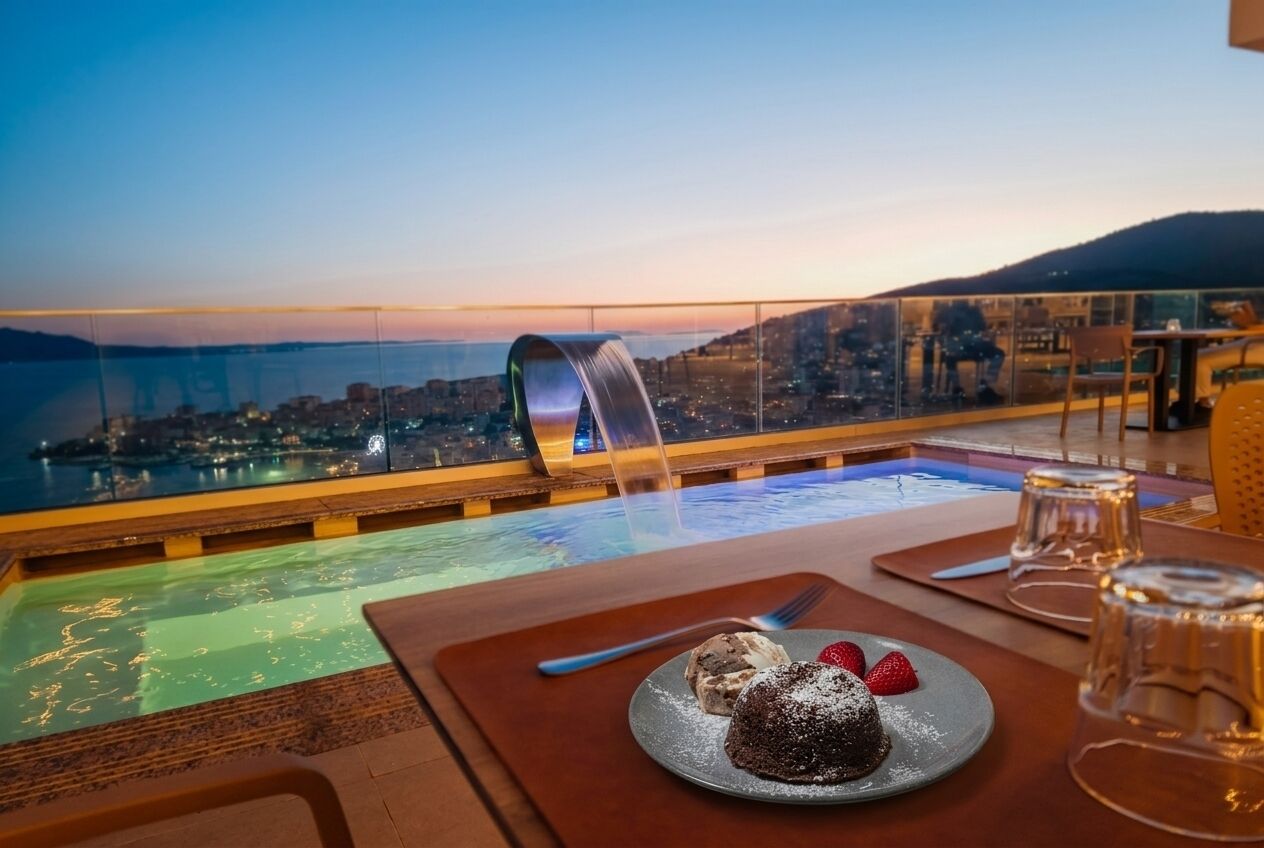 A dining table with a dessert, a glass of water, and a view of a cityscape at dusk.
