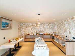 A modern kitchen with a stone wall backsplash, wooden cabinets, and a long dining table set for a meal. - Sunset Shoreline Saranda Hotel (Sarandë)