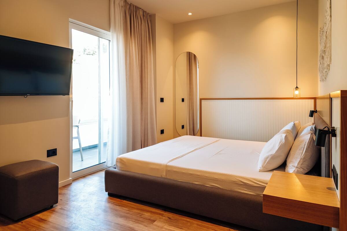 A modern bedroom with a large bed, a wooden bench, a TV mounted on the wall, and a sliding glass door leading to a balcony.