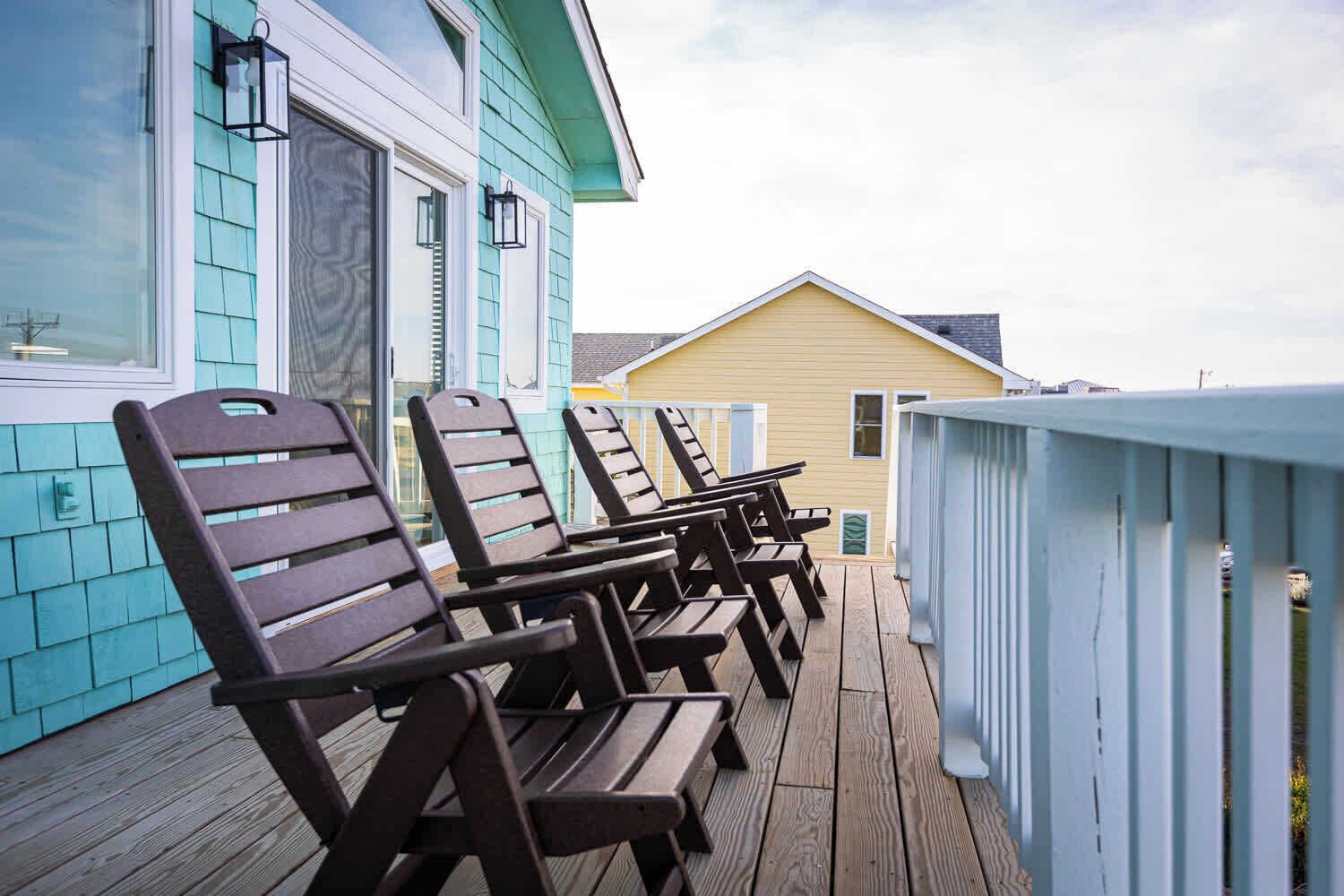 A wooden deck with lounge chairs and a railing, adjacent to a building with teal siding and a yellow roof.
