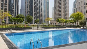 Una piscina amb baranes metàl·liques, envoltada per un solàrium pavimentat. Al fons, edificis residencials alts amb façanes de vidre reflectant.