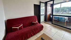 A red sofa with cushions, a small wooden table with a remote control, and a large sliding glass door leading to an outdoor area with chairs. - Cozy 1BR Apt with Terrace, 4 Sleepers, Near Beach and Port, Parking Included (Cap d'Agde)