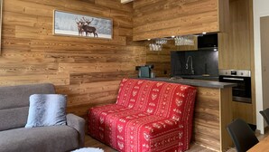 A modern kitchen with wooden paneling, a dining area with a red patterned sofa, and a framed deer picture on the wall. - Charming renovated studio with balcony and WIFI, near the slopes (Pra-Loup)