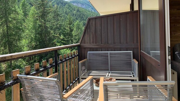 A balcony with a view of a forested mountain, a wooden railing, and a covered seating area with metal chairs. - Charming renovated studio with balcony and WIFI, near the slopes (Pra-Loup)