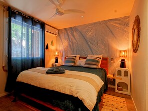 A bedroom with a bed, a ceiling fan, a wall-mounted air conditioner, and a bedside table with a lamp.
