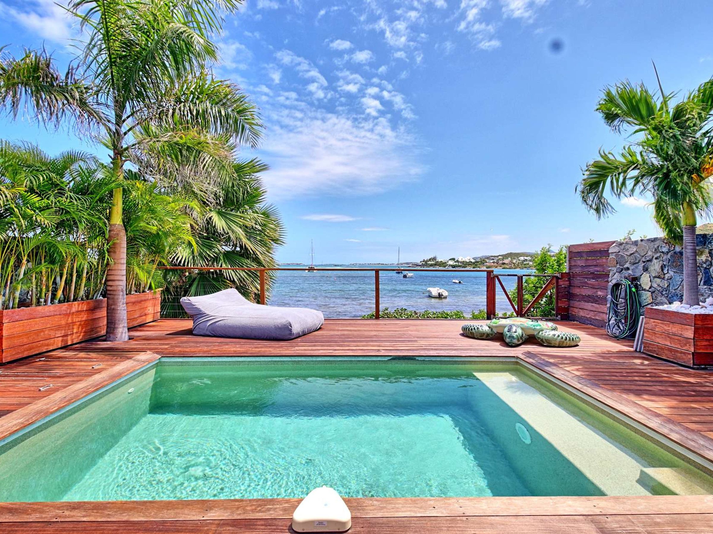 A wooden deck with a pool, palm trees, and a view of the sea.