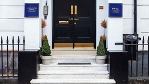 A black double door with a brass handle, flanked by two potted plants and a staircase leading up. - Putera Puteri Residence (London)