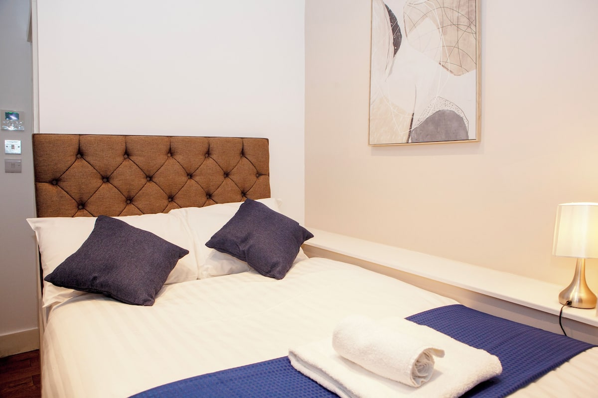 Una cama bien tendida con un cabecero capitonado, dos almohadas y una toalla enrollada sobre un tapete azul.