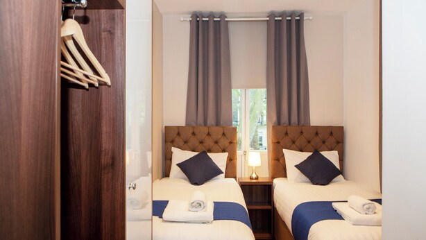 A hotel room with two beds, a wooden wardrobe, a window with curtains, and a nightstand with a lamp. - Putera Puteri Residence (London)