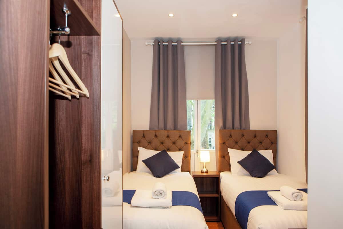 Habitación de hotel con dos camas, un armario de madera, una ventana con cortinas y una mesita de noche con lámpara.