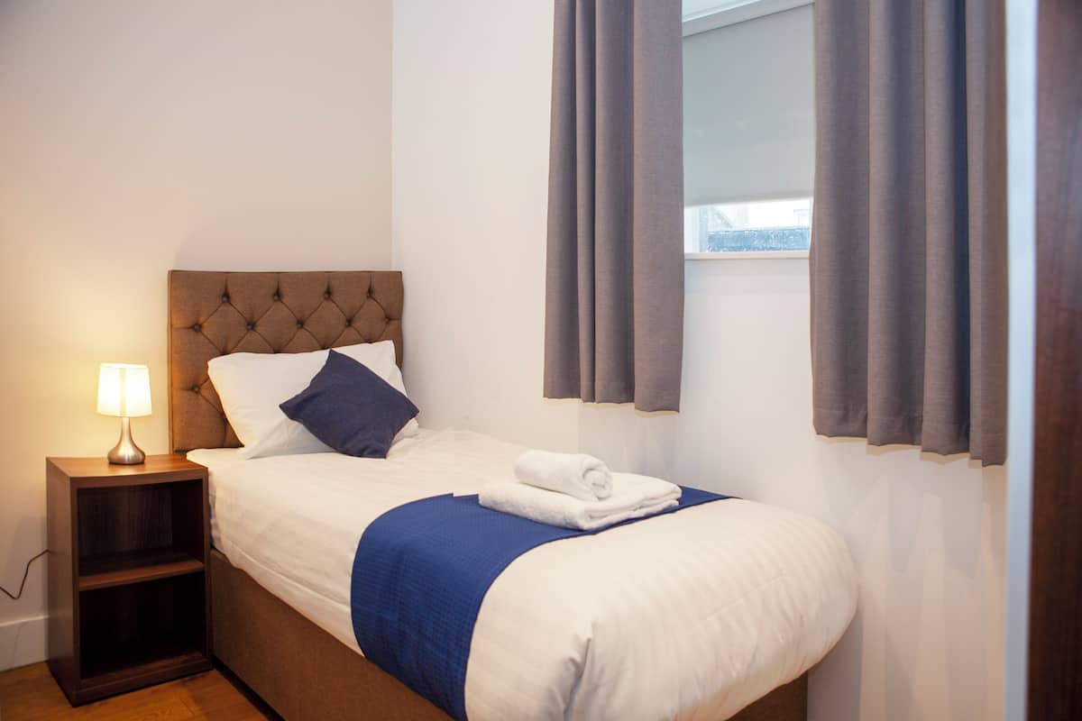 Una cama bien hecha con una manta azul, una mesita de noche de madera con una lámpara y una ventana con cortinas.