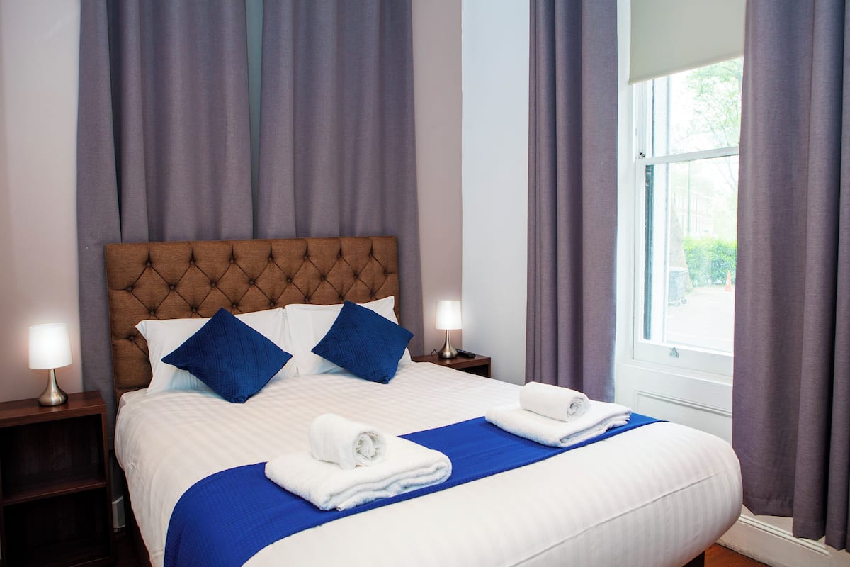 Una cama bien hecha con cabecera capitonada, dos almohadas azules y dos toallas blancas.